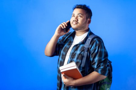 Téléchargez les photos : Un étudiant adulte tenant des livres et parlant sur un téléphone intelligent, isolé en fond bleu - en image libre de droit