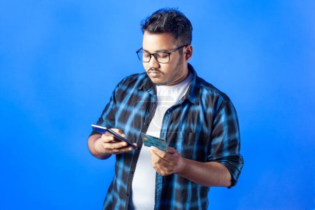 Téléchargez les photos : Plan recadré d'un étudiant masculin utilisant un téléphone portable avec carte de crédit, isolé sur fond bleu - en image libre de droit