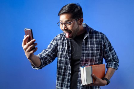 Photo for In single color background a university male student with wide eyes showing tongue out when doing video call on phone. - Royalty Free Image