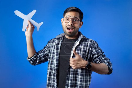 Photo for Indian ethnicity, toothy smile, journey, airplane. Man with thumb up hand gesture showing paper cut airplane. - Royalty Free Image
