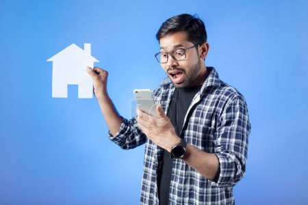 Photo for House rental, home insurance, Indian ethnicity. A shocked college student looking at phone and holding paper cutting airplane. - Royalty Free Image