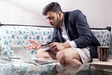 Téléchargez les photos : Homme indien avec expression haussant les épaules tout en utilisant la carte de crédit pour le paiement des achats en ligne pendant la quarantaine à domicile - en image libre de droit