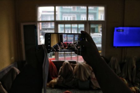 Foto de Noviembre 01,2023, Srinagar Cachemira, India: Una niña toma una foto de su teléfono mientras las novias con velos se sientan en el suelo mientras esperan asistir a una ceremonia de matrimonio en masa, en Srinagar - Imagen libre de derechos