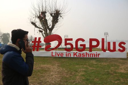 Téléchargez les photos : 12 décembre 2023, Srinagar Cachemire, Inde : Un homme parle sur son téléphone portable passé # Airtel5Gplus est vu sur les rives du lac Dal lors de la 5G en direct plus l'expérience à Srinagar. Airtel a annoncé le lancement de ses services 5G de pointe - en image libre de droit