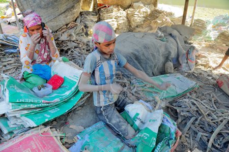 Foto de 19 diciembre 2023 Sylhet-Bangladesh: Un niño pequeño con su madre trabajando por un ingreso extra para la familia está arriesgando sus vidas trabajando en el polvo de las bolsas de cemento junto al río Surma en Sylhet, Bangladesh - Imagen libre de derechos