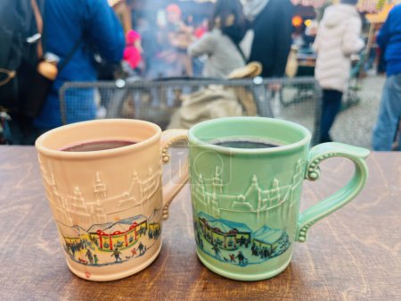 Photo for Two beautiful colorful mulled wine mugs with mulled wine at a Christmas fair. The mugs have a picture of the city. Green mug. Brown mug. A bonfire with firewood in the background. - Royalty Free Image