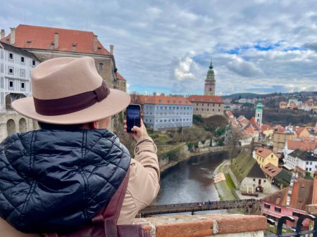 Female tourist in a hat, taking photos of Cesky Krumlov with her mobile phone. An old fairy tale town. Magical atmosphere and magnificent sky. High quality photo.