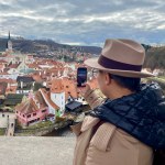Female tourist in a hat, taking photos of Cesky Krumlov with her mobile phone. An old fairy tale town. Magical atmosphere and magnificent sky. High quality photo. 