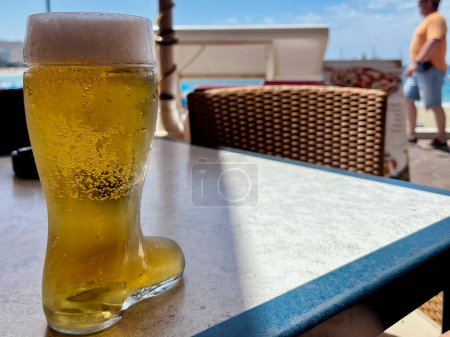 Boot shaped beer glass with frothy golden lager drink on table at outdoor cafe on sky background. High quality photo