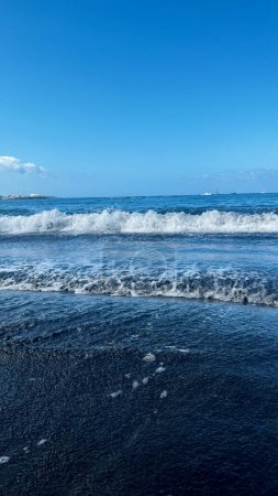 Atlantische Wellen treffen den Sandstrand. Schwarzer Sand und blauer Himmel. Vertikales Bild. Costa Adeje, Spanien. Hochwertiges Foto