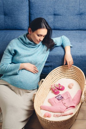 Téléchargez les photos : Happy mother enjoying pregnancy with wicker basket of cute tiny stuff newborn. Beautiful pregnant woman at home. Pregnant woman preparing for baby birth her daughter. Last months of pregnancy. - en image libre de droit