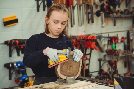 Téléchargez les photos : Cute boy makes wooden clock in the workshop. Young carpenter working with wood and sandpaper in craft workshop. School, development and learning concept. - en image libre de droit