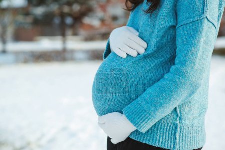Téléchargez les photos : Pregnant woman in blue sweater touching big belly with hands. Warm clothes in cold weather. Pregnancy time. Baby expectation. Pregnant woman's belly over winter background. - en image libre de droit