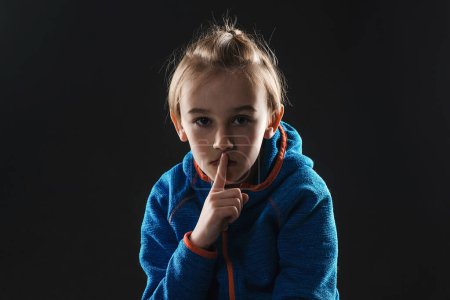 Foto de Young boy suffering from anxiety. Stressed alone boy over black background. Unhappy young boy with a finger on lips. Sad afraid child sitting quiet in dark. - Imagen libre de derechos