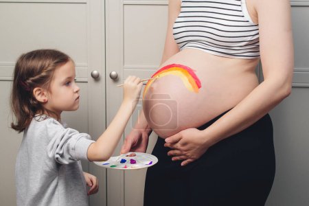 Photo for Happy little girl drawing rainbow on pregnant belly her mother. Baby birth expecting time and belly painting. Pregnant mom and child having fun together at home. - Royalty Free Image