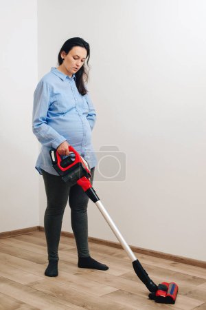 Foto de Mujer embarazada limpiando piso con aspiradora de mano. A la joven embarazada le gusta limpiar su casa. Fácil limpieza con aspiradora inalámbrica. Limpieza fácil moderna. - Imagen libre de derechos