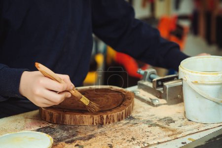 Téléchargez les photos : Boy is putting a protective mordant on the wooden disk, closeup. Young carpenter working with wood in craft workshop. Boy makes wooden clock in the workshop. - en image libre de droit