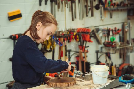 Photo for Cute boy makes wooden clock in the workshop. Young carpenter working with wood in craft workshop. School, development and learning concept. Artisan boy is putting a protective mordant on the wood. - Royalty Free Image
