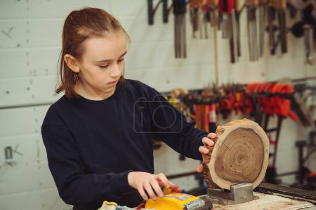 Téléchargez les photos : Young carpenter working with wood and sandpaper in craft workshop. School, development and learning concept. Cute boy makes wooden clock in the workshop. - en image libre de droit