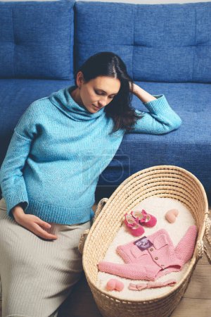 Téléchargez les photos : Pregnant woman preparing for baby birth her daughter. Last months of pregnancy. Happy mother enjoying pregnancy with wicker basket of cute tiny stuff newborn. Beautiful pregnant woman at home. - en image libre de droit