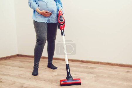 Téléchargez les photos : Young pregnant woman enjoys cleaning her house. Modern easy cleaning. Easy cleaning with a wireless vacuum cleaner. Pregnant woman cleaning floor with handheld vacuum cleaner. - en image libre de droit