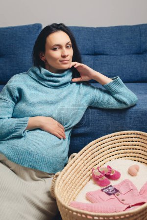 Foto de Pregnant woman preparing for baby birth her daughter. Last months of pregnancy. Happy mother enjoying pregnancy with wicker basket of cute tiny stuff newborn. Beautiful pregnant woman at home. - Imagen libre de derechos