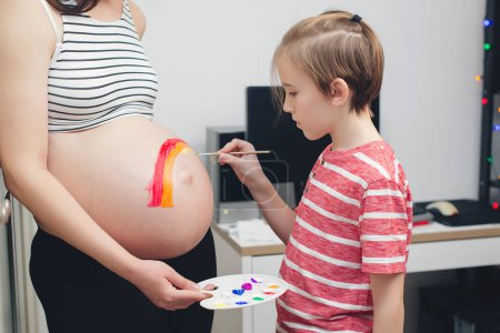 Photo for Cute boy drawing rainbow on pregnant belly his mother. Baby birth expecting time and belly painting. Pregnant mom and her son having fun together at home. Family, healthy pregnancy. - Royalty Free Image