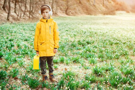 Téléchargez les photos : Joli garçon marchant sur la nature. Enfant avec casque relaxant dans le parc de printemps. Enfant élégant avec écouteurs dans le parc. - en image libre de droit