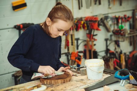 Photo for Artisan boy is putting a protective mordant on the wood. Cute boy makes wooden clock in the workshop. Young carpenter working with wood in craft workshop. School, development and learning concept. - Royalty Free Image