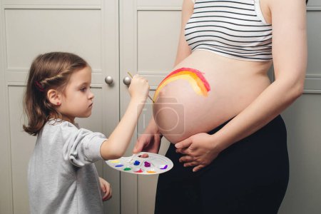 Téléchargez les photos : Happy little girl drawing rainbow on pregnant belly her mother. Baby birth expecting time and belly painting. Pregnant mom and child having fun together at home. - en image libre de droit
