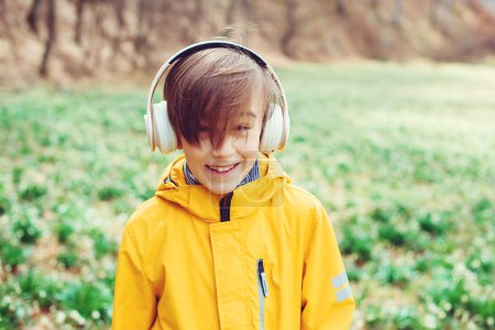Téléchargez les photos : Joyeux garçon écoutant de la musique à la nature. Enfant avec casque relaxant dans le parc de printemps. Garçon souriant avec écouteurs sur la promenade. Mode de vie, enfance et émotions. - en image libre de droit