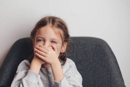 Photo for Little girl covers mouth with hand. Sad offended child girl sitting at sofa at home. Child abuse. Violence, domestic problem in family. - Royalty Free Image