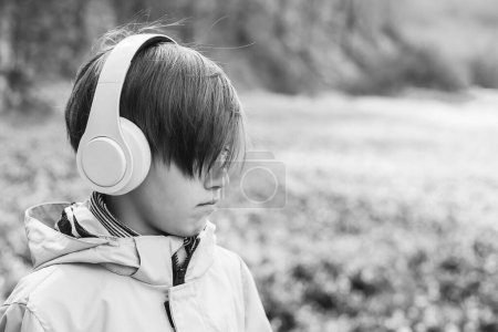 Photo for Upset boy with headphones on the walk. Lifestyle, childhood and emotions. Sad boy listening to music at nature. Kid with headphones relaxing in the spring park. - Royalty Free Image
