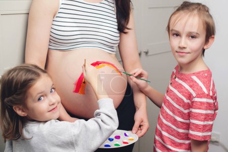 Téléchargez les photos : Baby birth expecting time and belly painting. Happy children and pregnant mom having fun together at home. Family, healthy pregnancy and baby birth. Happy kids painting pregnant belly their mother. - en image libre de droit
