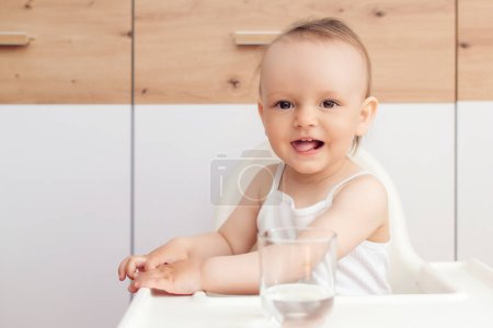 Photo for Sweet baby girl holding glass and drinking water. Happy baby sitting in a baby chair in the kitchen. Child drinks water. - Royalty Free Image