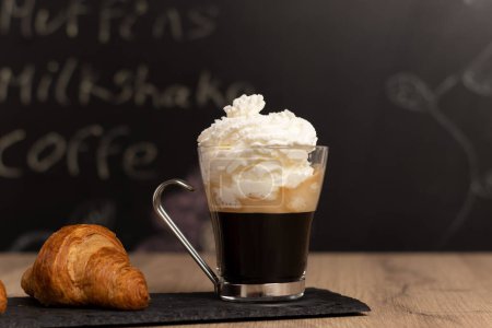 Photo for Coffee with cream, croissant, in slate stone, glass race with silver gray trim a wooden table and slate background written and out of focus with space for text. - Royalty Free Image