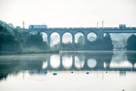 Photo for A goods train passes over the viaduct at Obersee in Bielefeld and the scene is reflected in the lake - Royalty Free Image