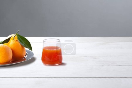 Photo for Orange Juice Drink in Glass on White Wooden Table and Gray Background  Whole Oranges on Silver Plate, Green Leaves, with Shadows, Red Juice  High Quality Minimalist Ingredient Photo - Royalty Free Image