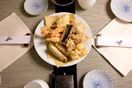 Foto de Tempura en plato de restaurante de comida japonesa con verduras y camarones fritos en masa, palillos, placas de cerámica, cena gourmet en mesa de madera negra y manteles de mesa Vista superior de alta resolución - Imagen libre de derechos