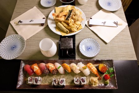 Photo for Sushi and Tempura in Japanese Food Restaurant  Gourmet Dishes, Ceramic Plates with Deep Fried Vegetables, Rows of Rolls and Nigiri, Red Caviar, Chopsticks, Black Wood Table and Placemats  Top View - Royalty Free Image
