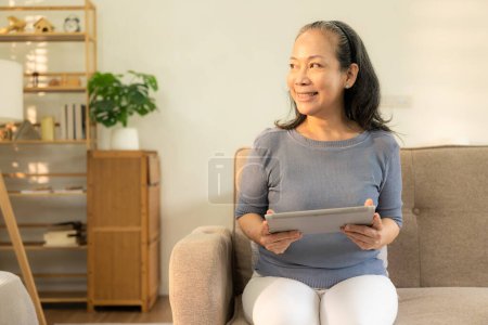 Téléchargez les photos : Heureuse femme asiatique âgée assise sur un canapé tenant une tablette numérique dans la main dans le salon Femme âgée utilisant des appareils intelligents informatiques à la maison utiliser la technologie de surf Internet - en image libre de droit