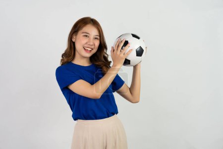 Téléchargez les photos : Heureuse asiatique fan de football envoyant un soutien à l'équipe préférée avec ballon de football, femme souriante en t-shirt bleu tenant ballon de football pour encourager au match de football, isolé sur fond blanc. - en image libre de droit