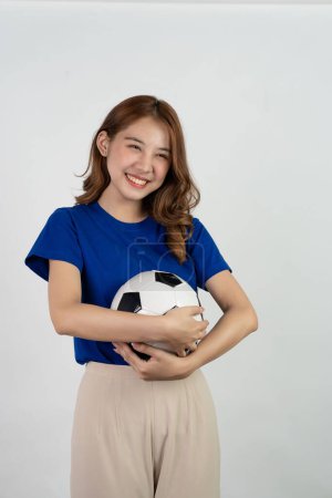 Foto de Feliz asiática fanática del fútbol enviando apoyo al equipo favorito con pelota de fútbol, mujer sonriente en camiseta azul sosteniendo pelota de fútbol para animar en el partido de fútbol, aislado sobre fondo blanco. - Imagen libre de derechos