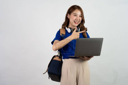 Foto de Feliz joven asiática estudiante con mochila de pie y la celebración de la computadora portátil. Mujer en camiseta azul trabajando en ordenador portátil aislado sobre fondo blanco. Concepto de educación en línea. - Imagen libre de derechos