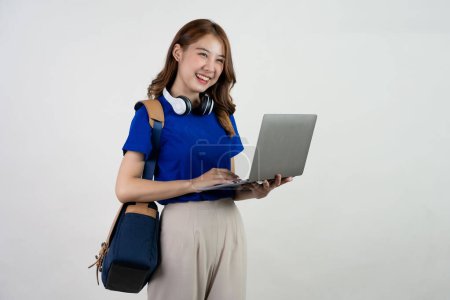 Foto de Feliz joven asiática estudiante con mochila de pie y la celebración de la computadora portátil. Mujer en camiseta azul trabajando en ordenador portátil aislado sobre fondo blanco. Concepto de educación en línea. - Imagen libre de derechos