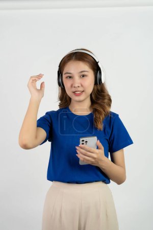 Téléchargez les photos : Jolie adolescente asiatique heureuse utilisant un téléphone mobile jouant de la musique à partir d'une application smartphone, souriant heureux tout en écoutant de la musique en streaming sur fond blanc, concept de technologie - en image libre de droit