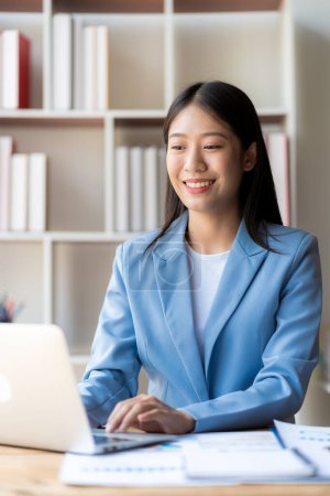 Téléchargez les photos : Heureuse jeune femme d'affaires asiatique travaillant avec des documents financiers et un ordinateur portable et un smartphone - en image libre de droit