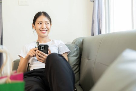 Photo for Asian woman holding credit card and smart phone and laptop sitting on sofa at home doing online banking transactions in online shopping - Royalty Free Image