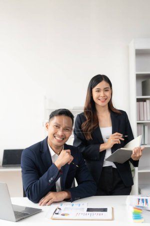 Photo for Two Asian business men and women smiling happily working with laptops and financial documents. They talk about their projects and brainstorm creative ideas together in the office. - Royalty Free Image