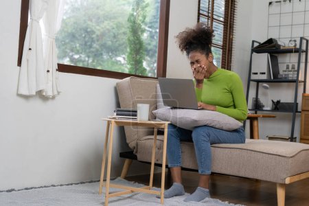 Foto de Atractiva joven africana que trabaja en la computadora portátil mientras está sentada en el sofá en casa con auriculares navegando por la red usando teléfono inteligente y tableta mientras se relaja en casa disfrutando del tiempo libre. - Imagen libre de derechos